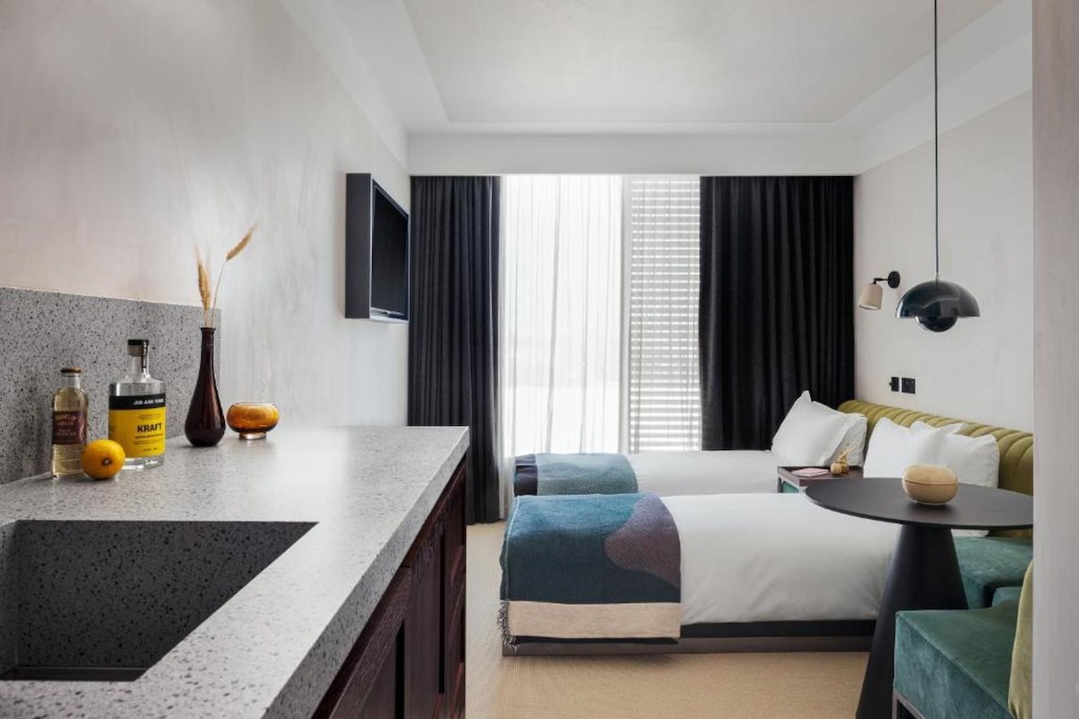 A hotel room with a bed, a desk, a chair, a small table, a TV, and a window with curtains.