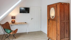Una habitació amb sostre inclinat, un armari de fusta amb mirall, un televisor fixat a la paret, una cadira amb coixí i una taula petita amb una làmpada.