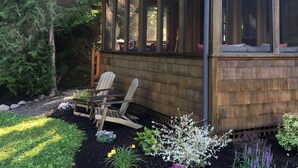 Una cabaña de madera con ventanas amplias, un porche cubierto y un jardín con diversas plantas.