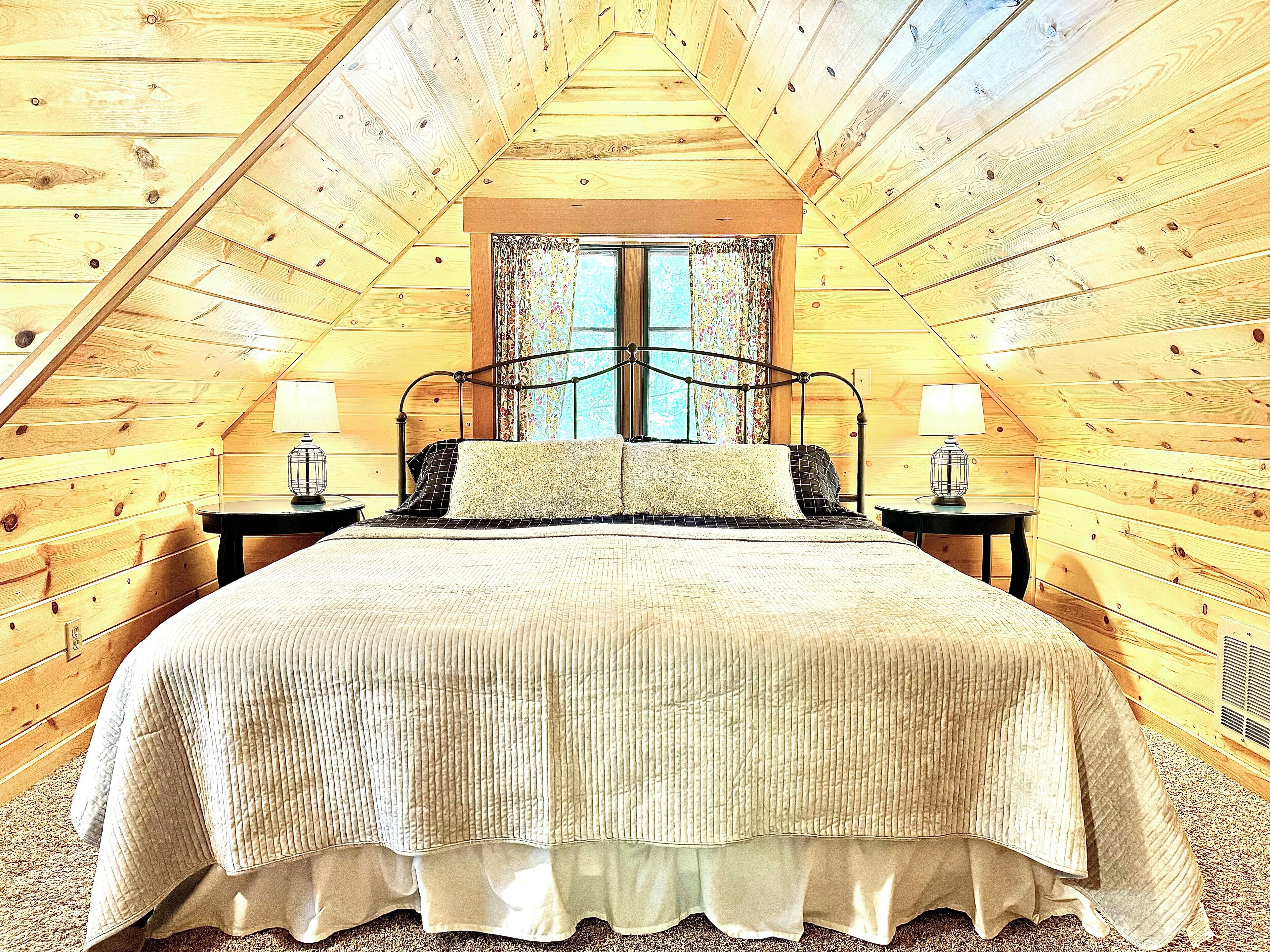 A cozy attic bedroom with a wooden sloped ceiling, a large bed with a quilted bedspread, two bedside lamps, and a window with floral curtains.