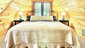 Un dormitorio acogedor en el ático con techo inclinado de madera, una cama grande con colcha acolchada, dos lámparas de noche y una ventana con cortinas estampadas.
