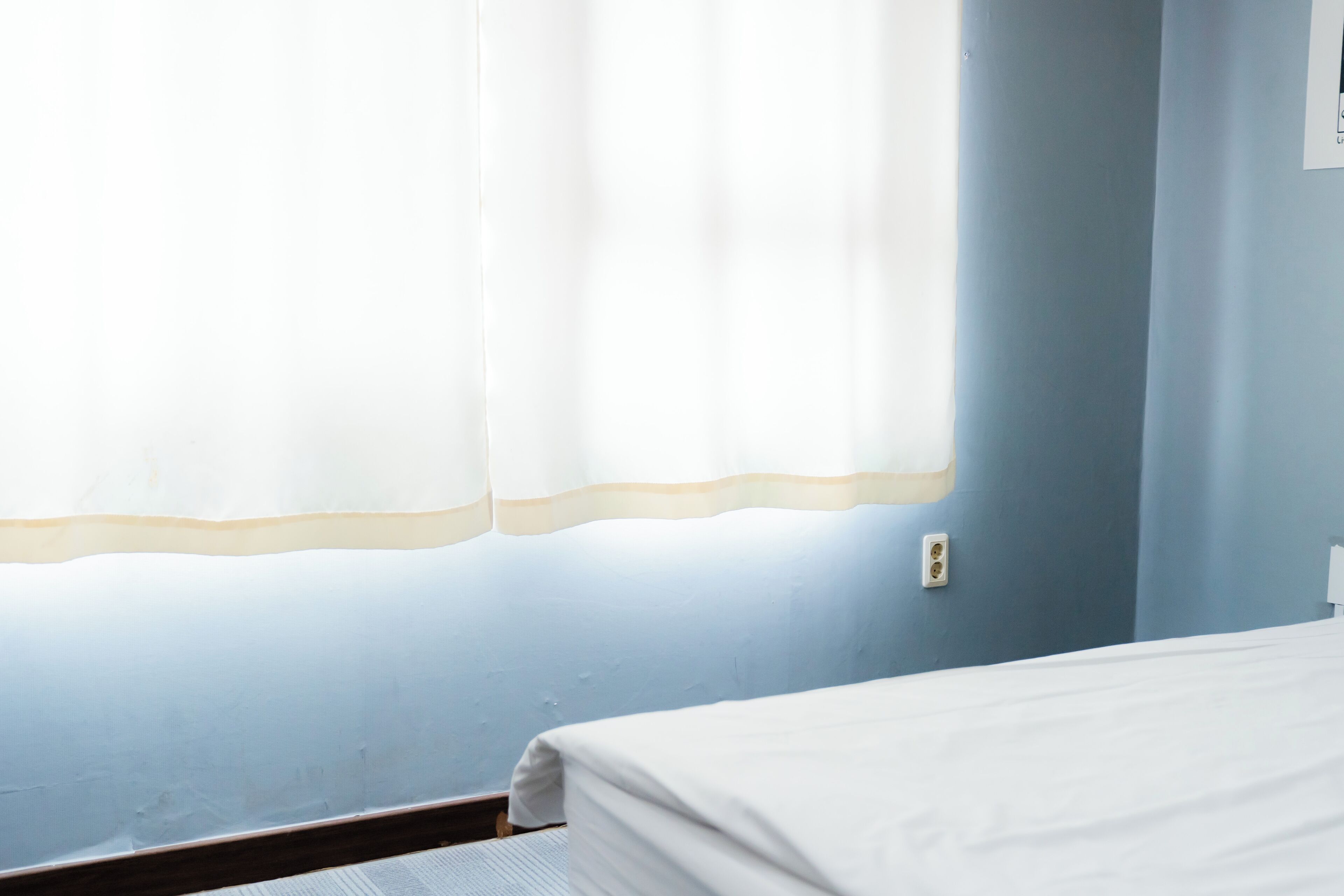 A room with a bed, a window with a white curtain, and a wall switch.