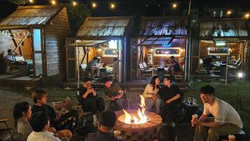 A group of people seated around a campfire in an outdoor setting with wooden structures and string lights.