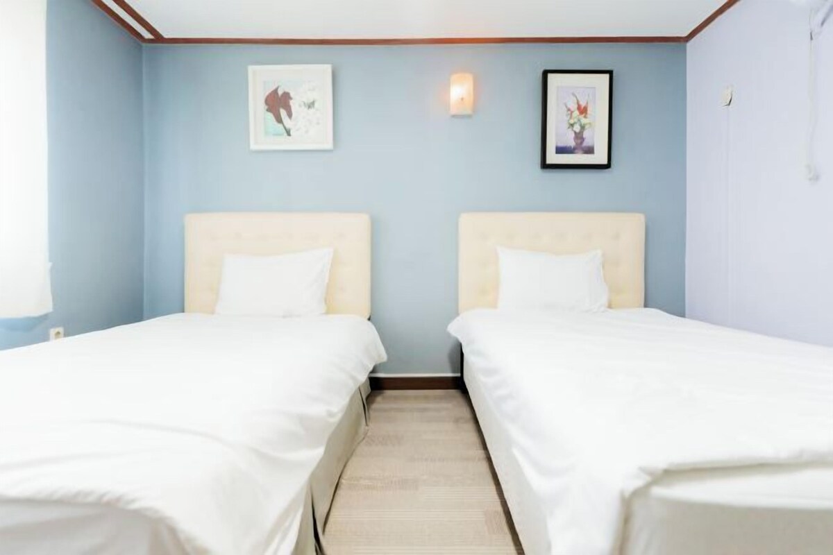 A hotel room with two single beds, a wooden floor, light blue walls, and two framed pictures on the wall.