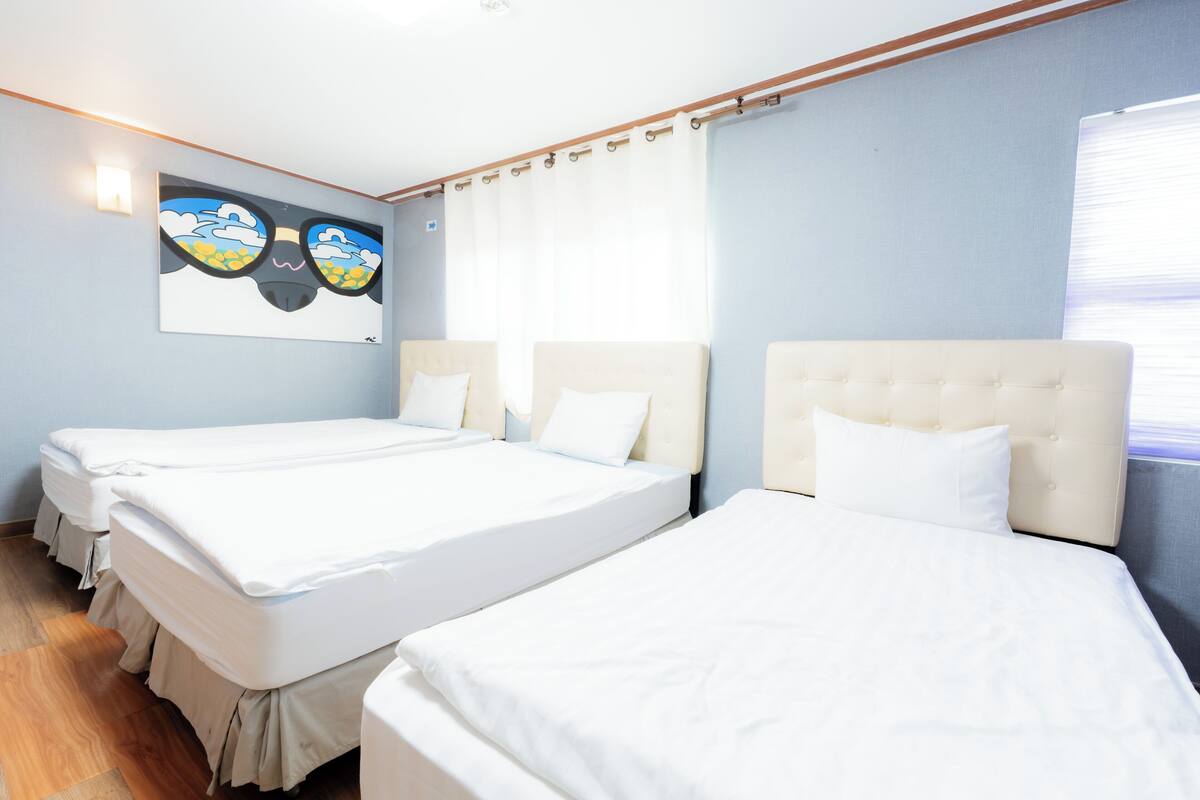 A hotel room with three beds, a painting on the wall, and a window with curtains.