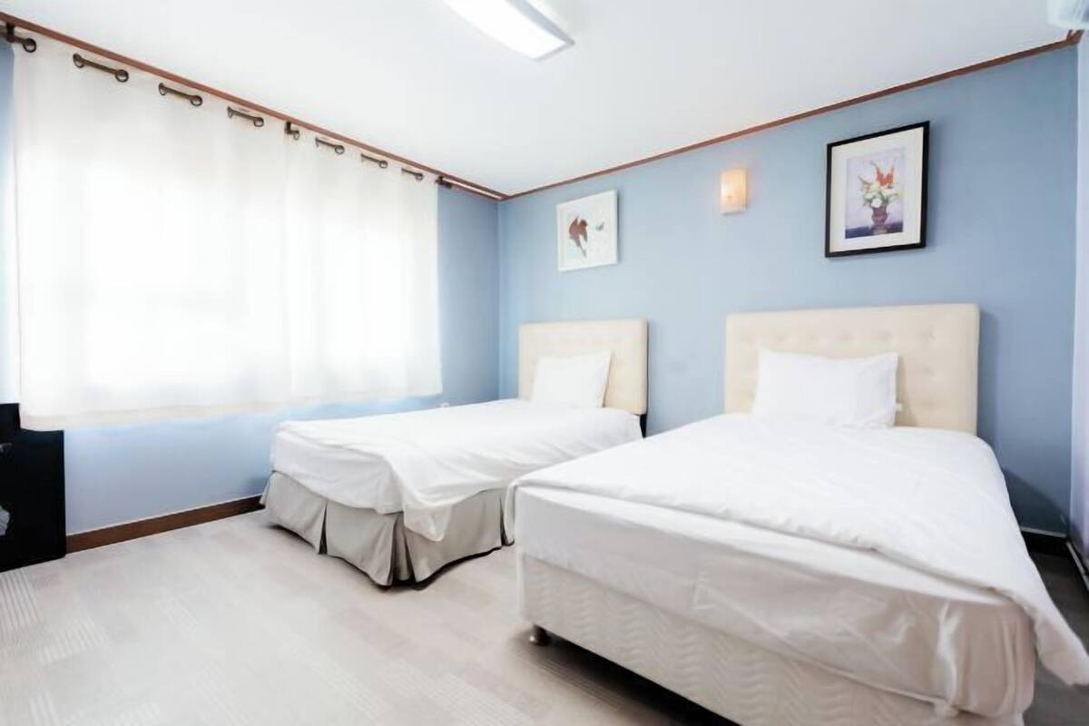 A hotel room with two beds, a large window with curtains, a framed picture on the wall, and a small table.