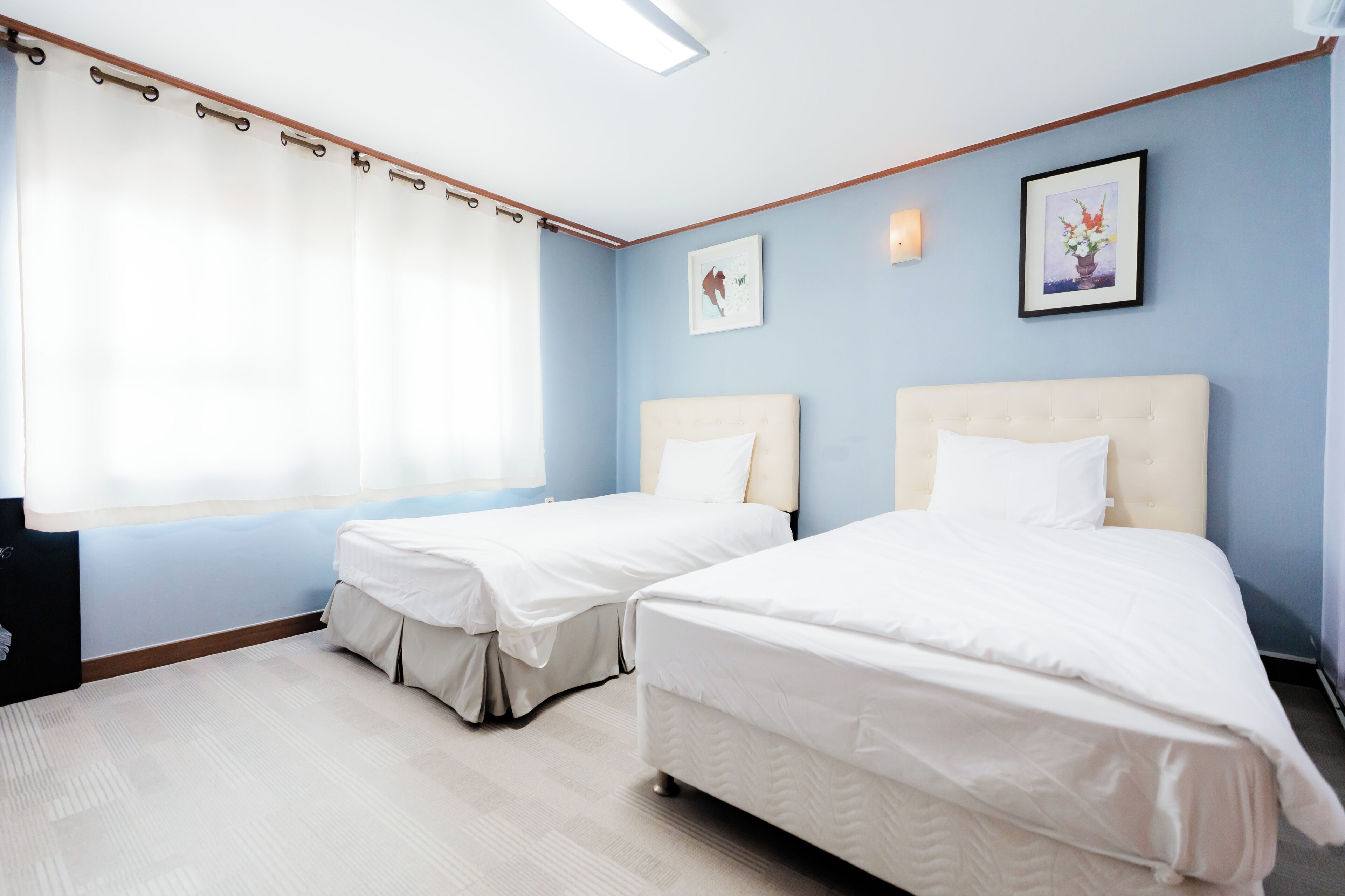 Una camera d'albergo con due letti, un'ampia finestra con tende, un quadro appeso al muro e un lampadario.