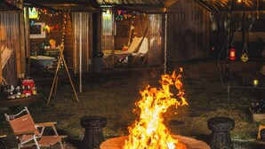 Een vuurkorf met rondom stoelen, een houten constructie op de achtergrond en lichtjes aan een draad.