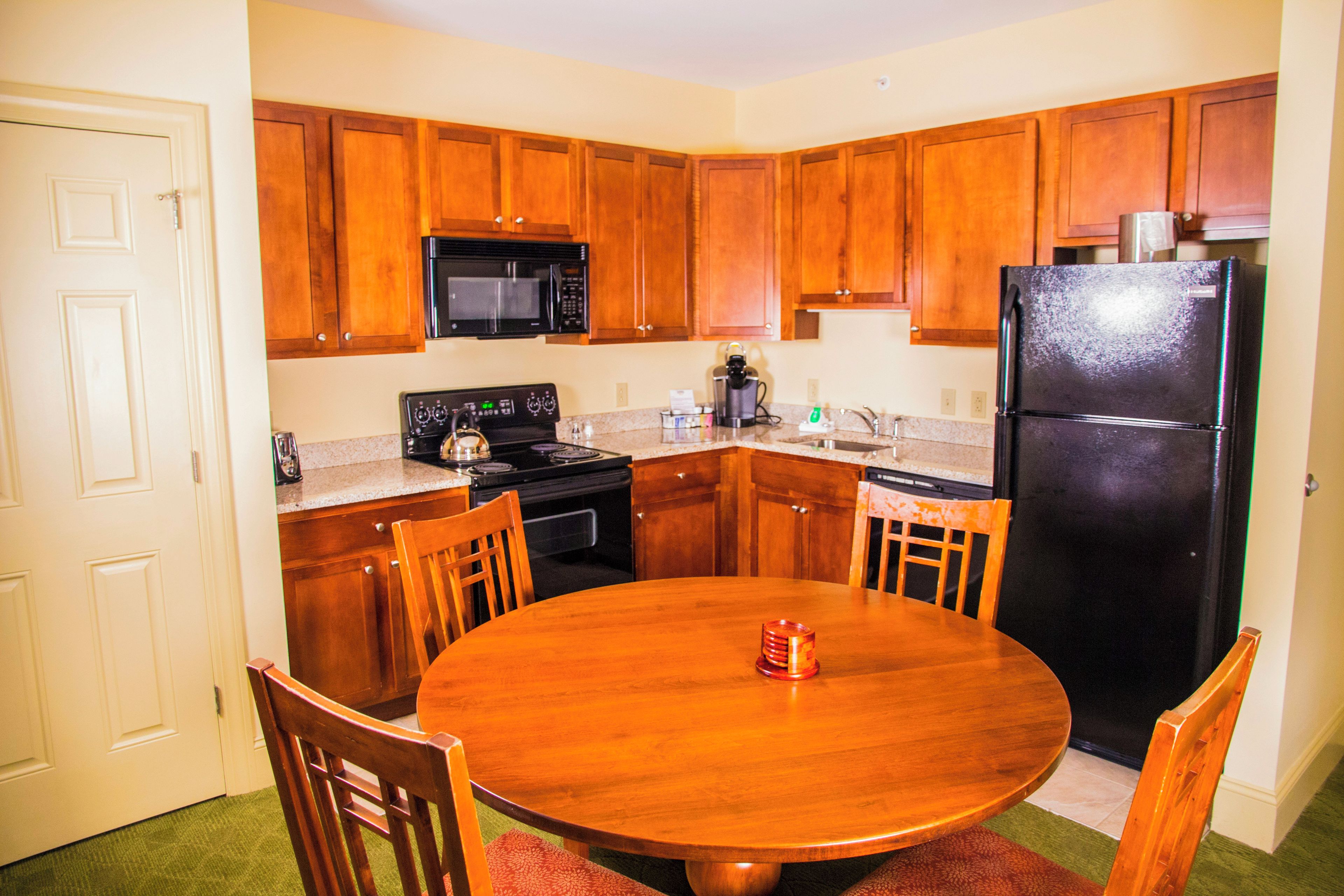 Cocina compacta con armarios de madera, un refrigerador negro, un microondas y una estufa. Hay una mesa redonda de madera con sillas.