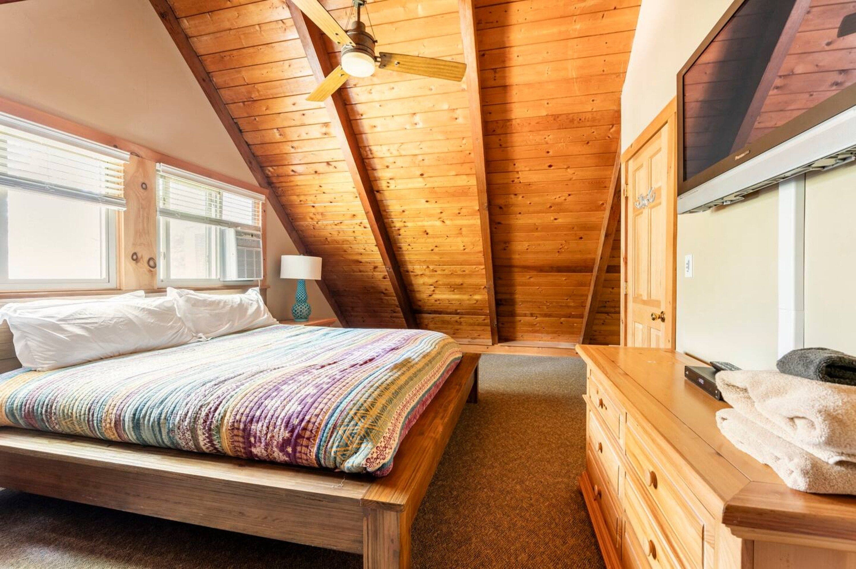 Un dormitorio con techo de madera, una cama con colcha colorida, un aparador de madera y un televisor en la pared.
