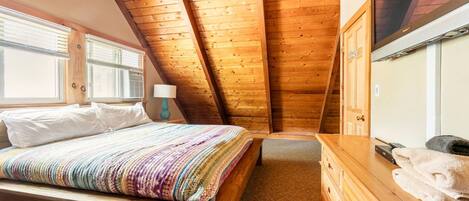 A bedroom with a wooden ceiling, a bed with a colorful blanket, a wooden dresser, and a TV on the wall.