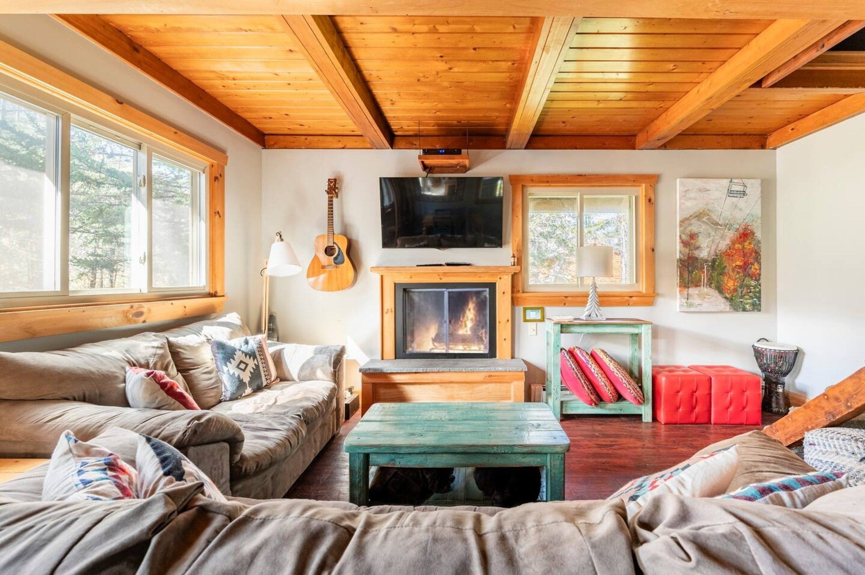 A cozy living room with a fireplace, wooden ceiling, and large windows.