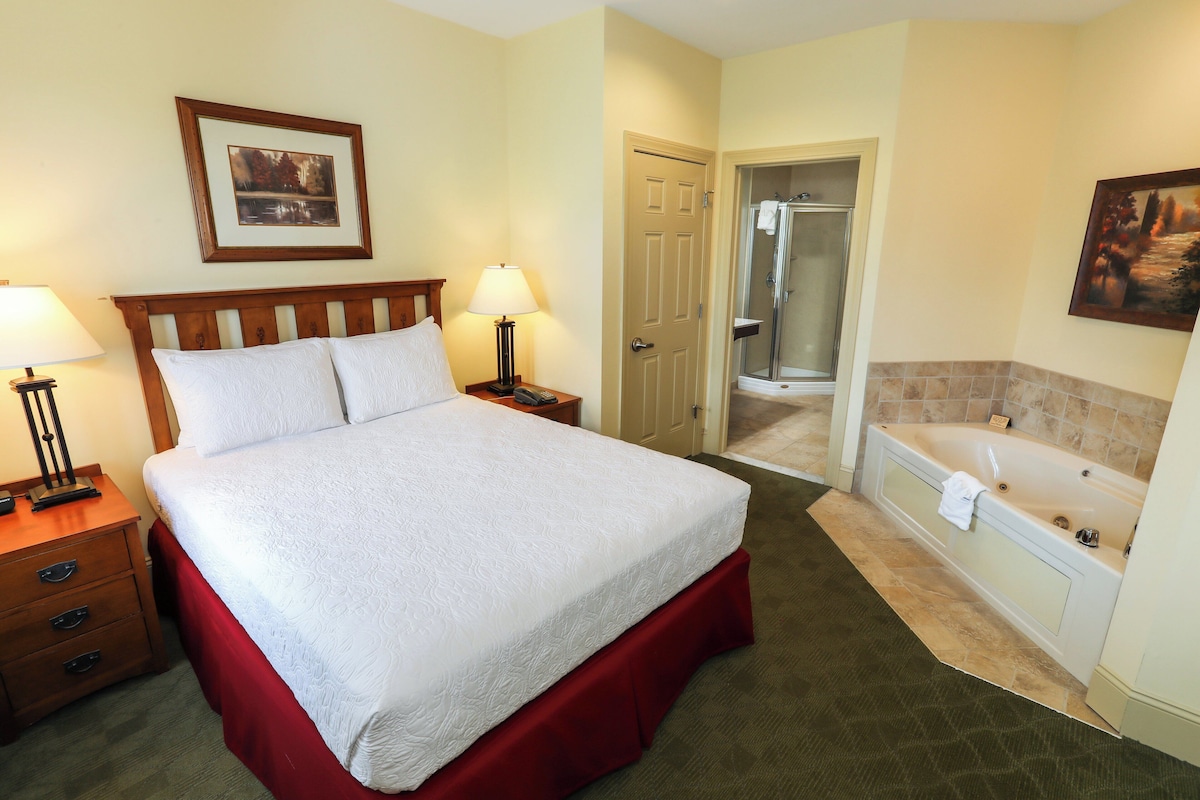 A hotel room with a large bed, a nightstand, a lamp, a bathroom with a bathtub and shower.
