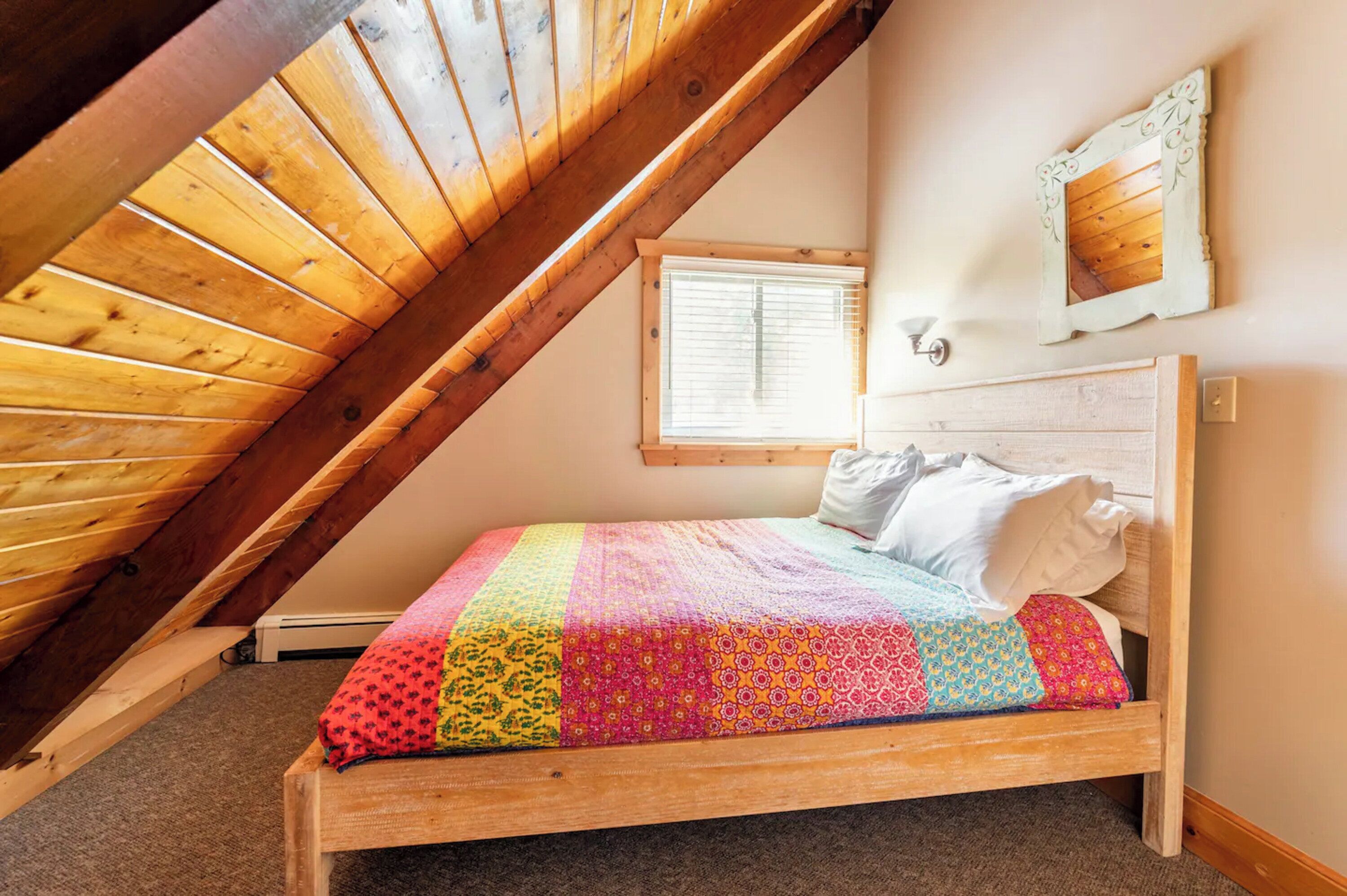 A bedroom with a sloped ceiling, a wooden bed frame, a colorful quilt, a window with blinds, and a wall-mounted light fixture.