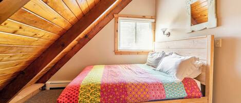 Un dormitorio con techo inclinado, estructura de cama de madera, colcha colorida, ventana con persianas y una lámpara de pared.