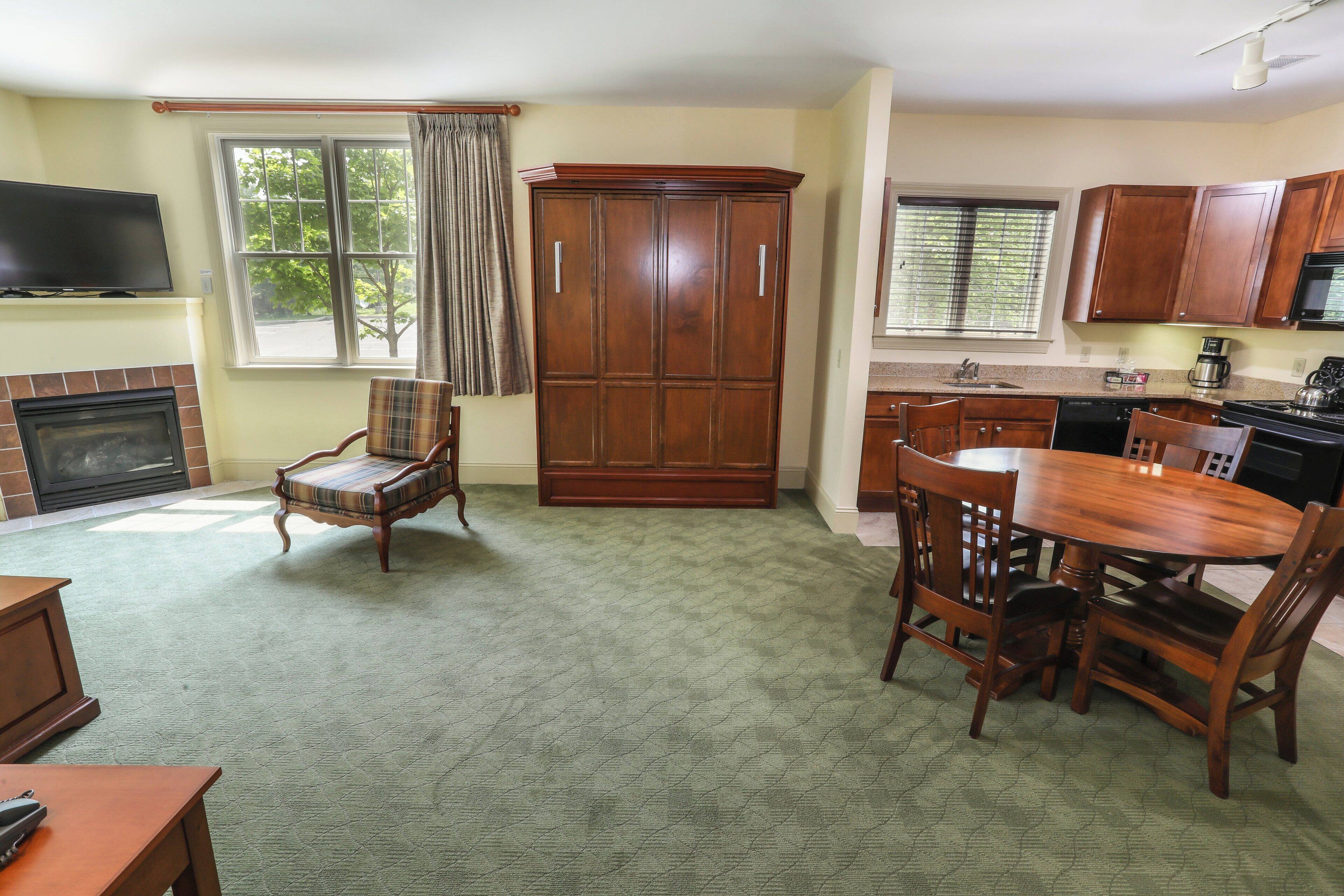 A living room with a fireplace, a television mounted above it, a plaid armchair, a wooden cabinet, and a dining area with a table and chairs.