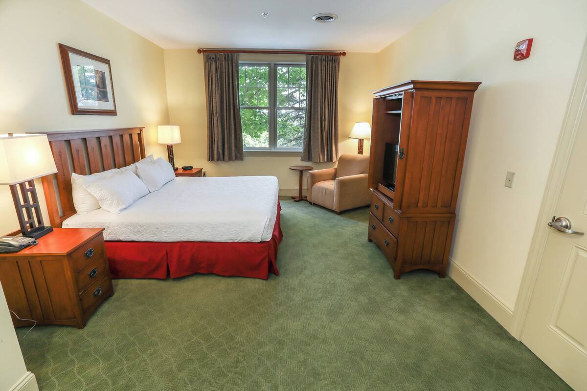 A hotel room with a large bed, a wooden wardrobe, a desk with a lamp, a chair, and a television.