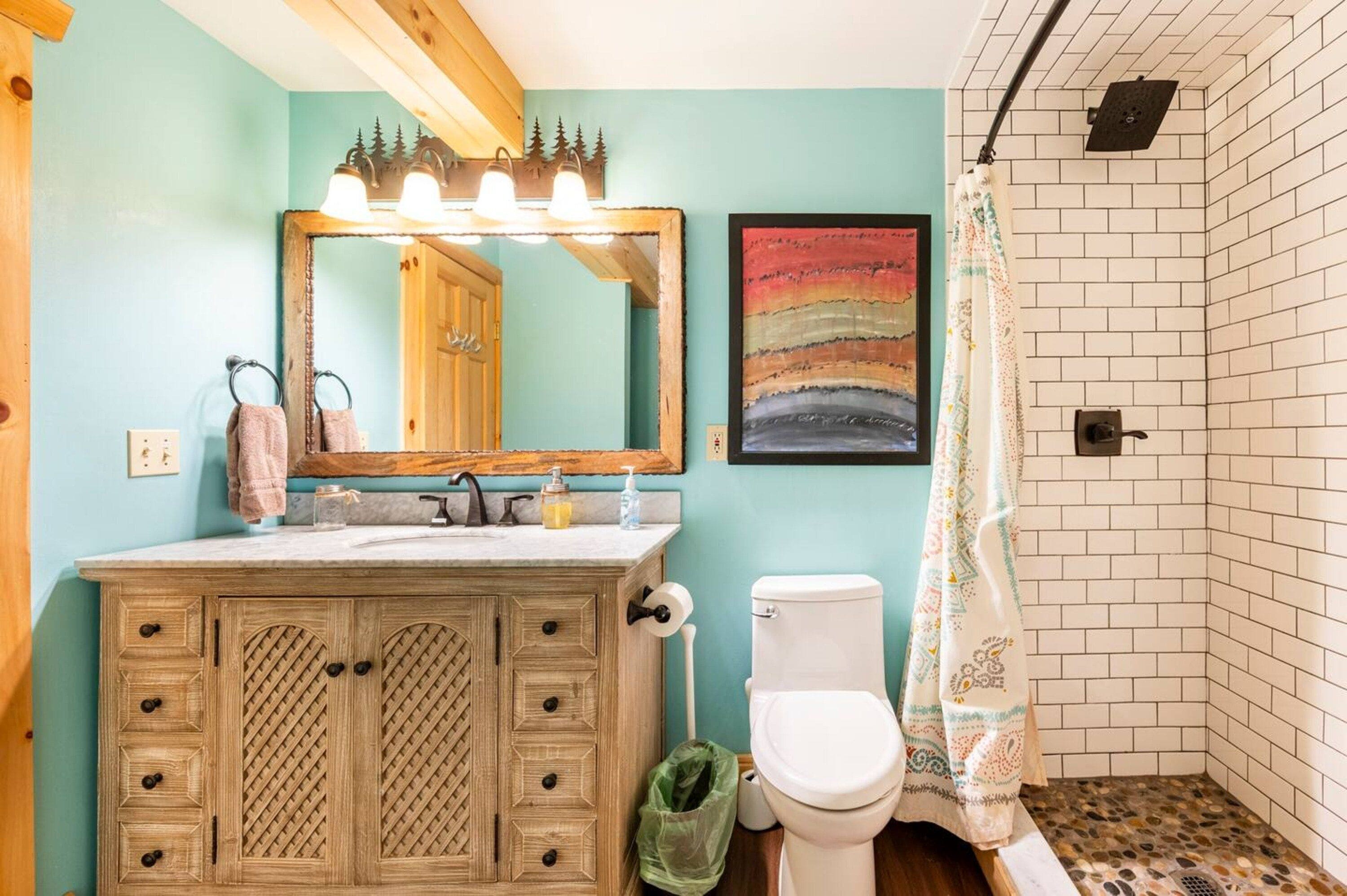 A bathroom with a wooden vanity, a white toilet, a shower with a patterned floor, and a mirror with lights.