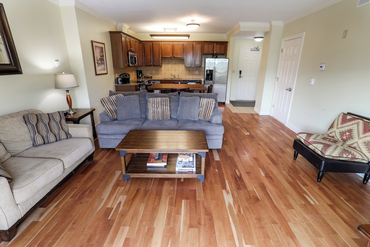 A living room with a sofa, a coffee table, and a chair. There is a kitchen in the background with wooden cabinets and appliances.