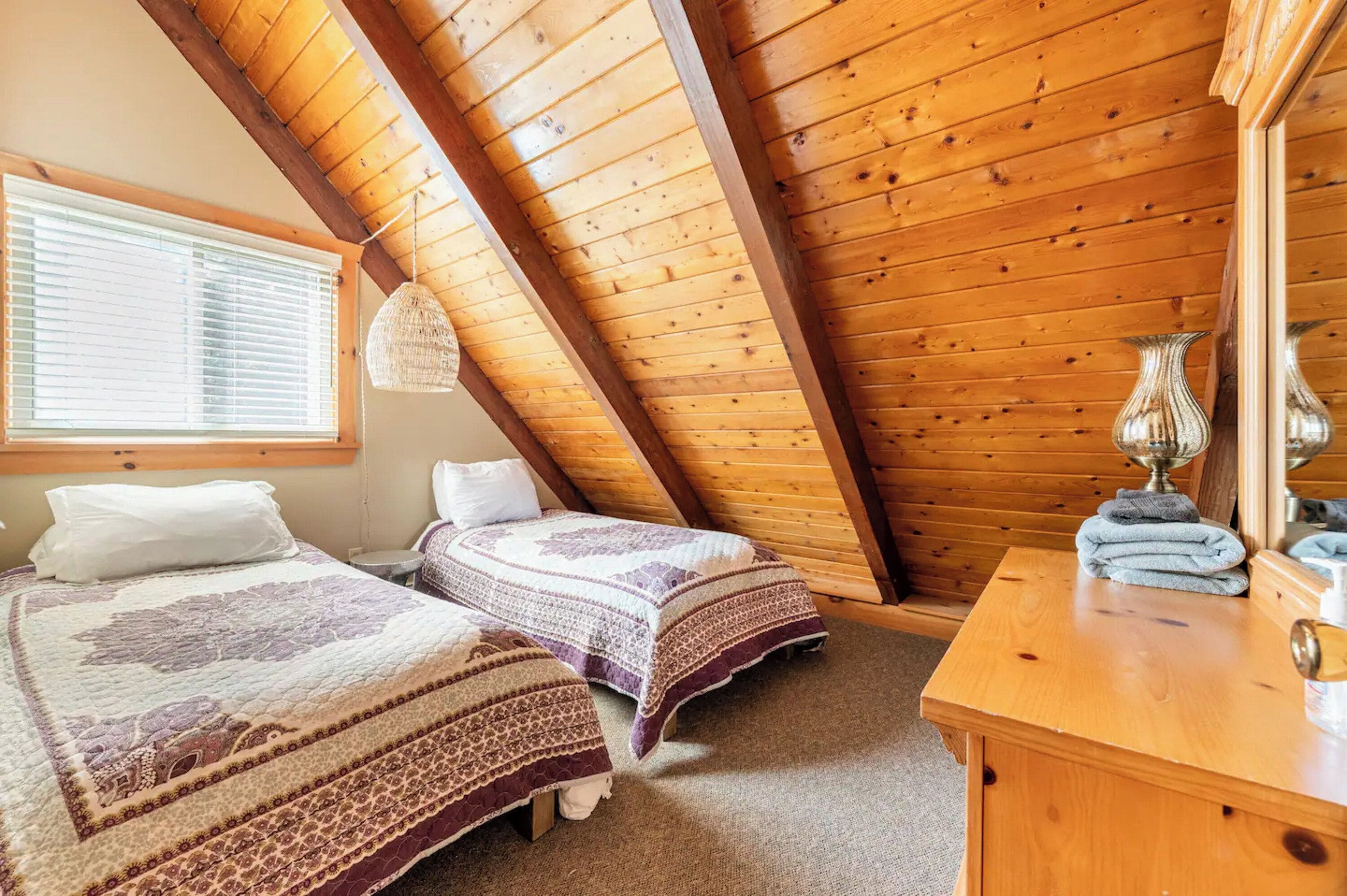Une chambre confortable avec deux lits, des poutres apparentes en bois au plafond, une grande fenêtre avec des stores et une commode en bois où sont rangées les serviettes.