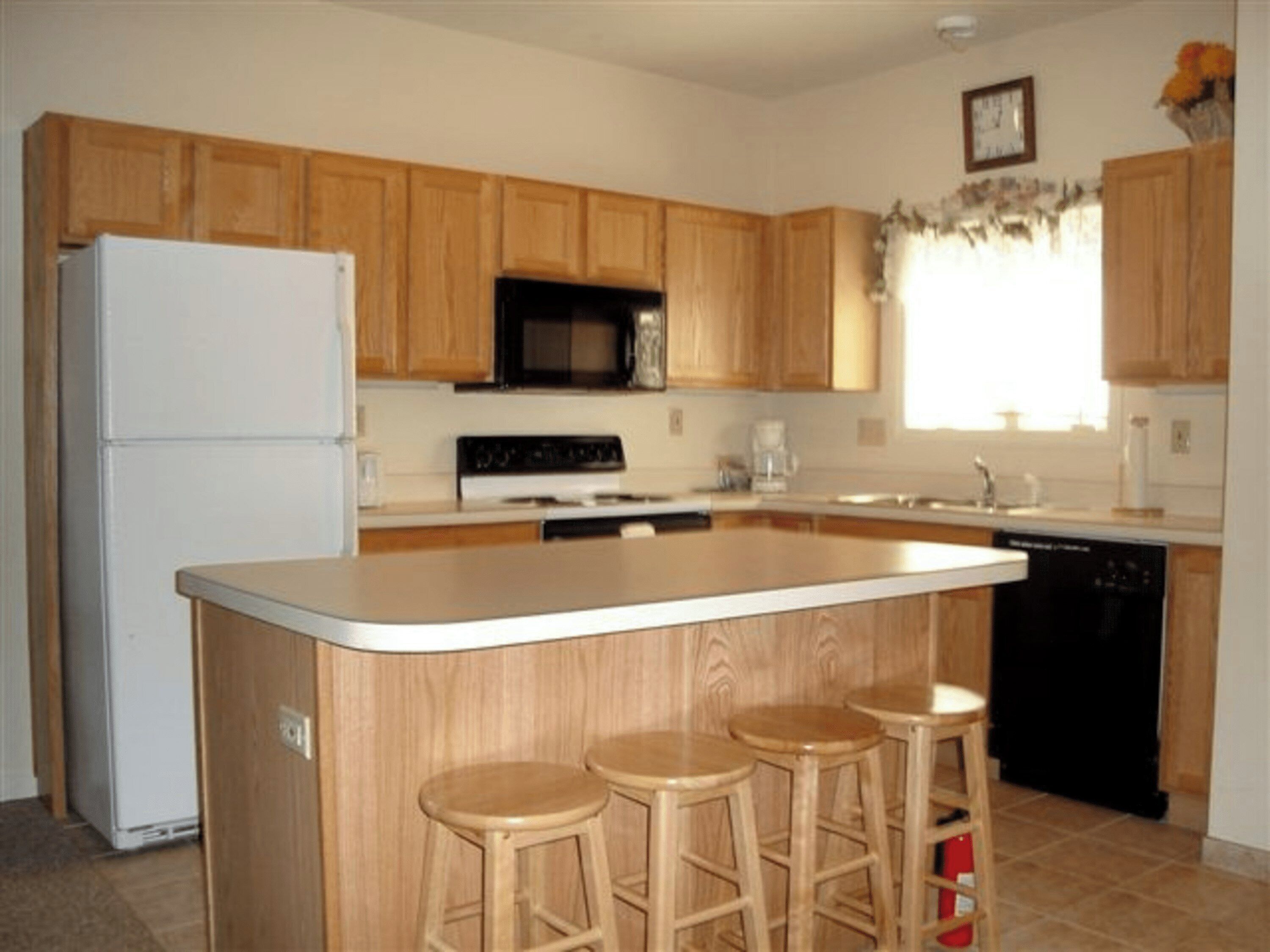 A kitchen with wooden cabinets, a white refrigerator, a black microwave, a stove, a sink, and three stools.