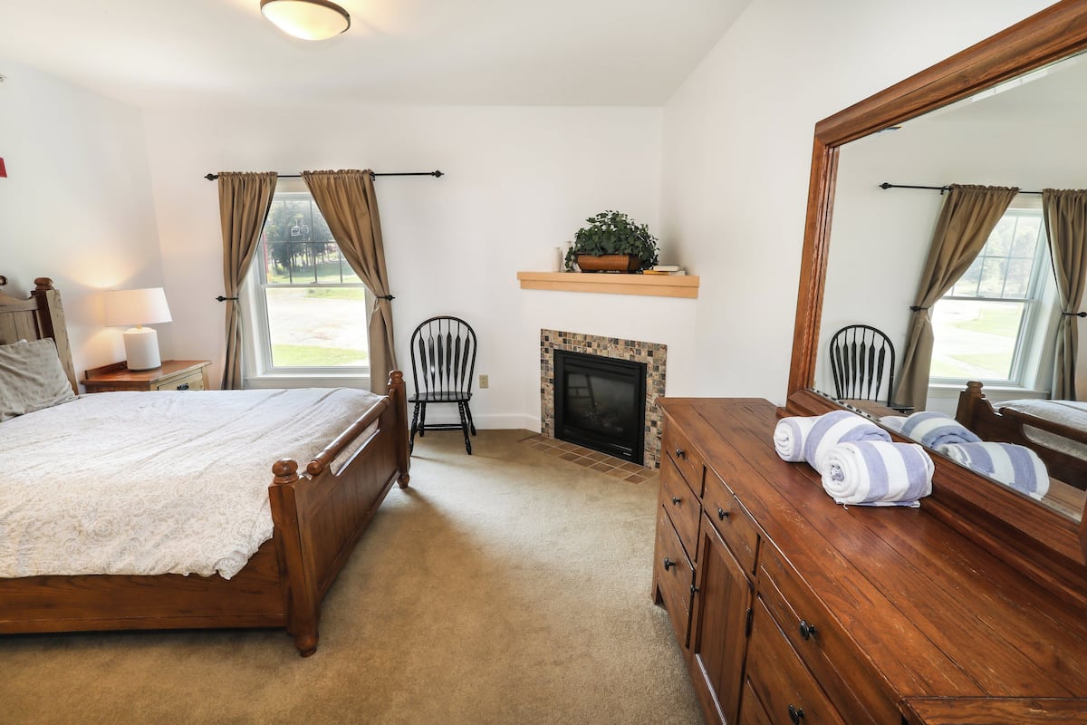 A bedroom with a wooden bed, a dresser, a mirror, a fireplace, and a window with curtains.
