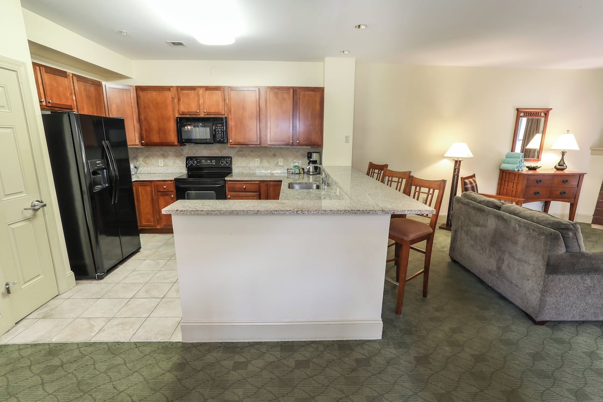 A modern kitchen with wooden cabinets, a black refrigerator, a microwave, and a coffee maker. There is a dining area with a sofa and chairs.