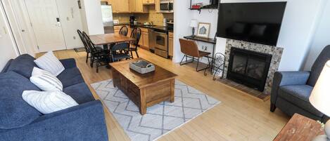 Ein modernes Wohnzimmer mit Sofa, Couchtisch und Kamin. Es gibt eine Essecke mit Stühlen und Tisch. Die Küche zeichnet sich durch Holzschränke und eine Mikrowelle aus.