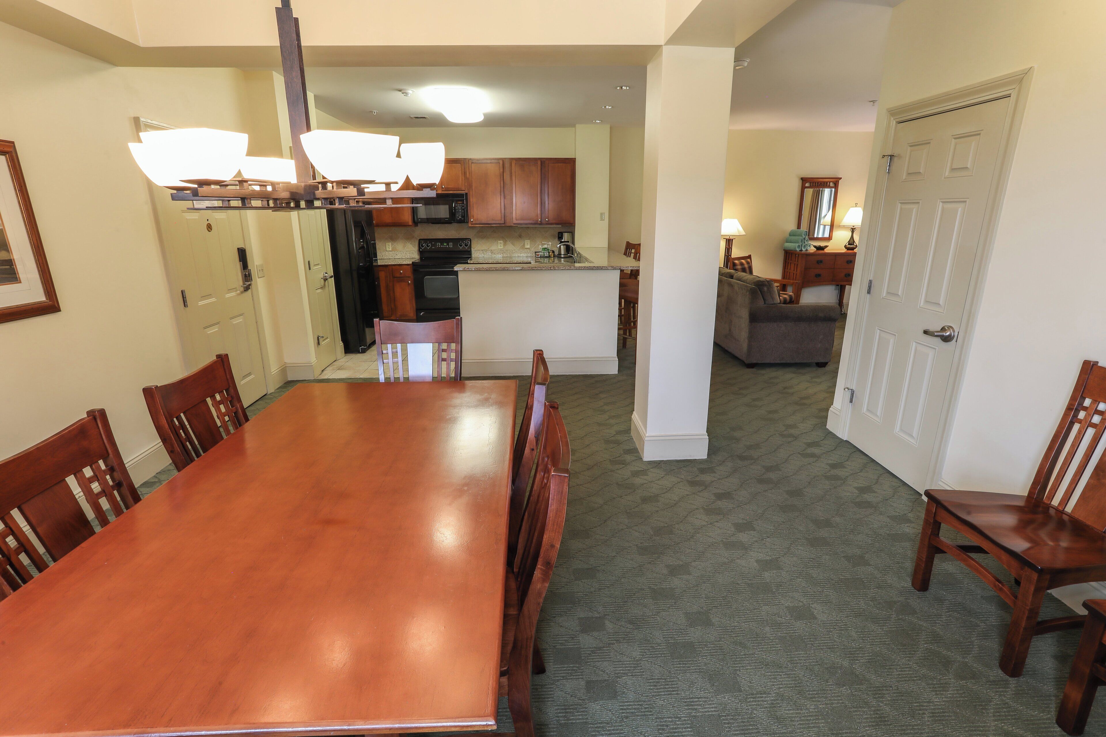 A dining area with a wooden table and chairs, a kitchen in the background, and a door leading to another room.