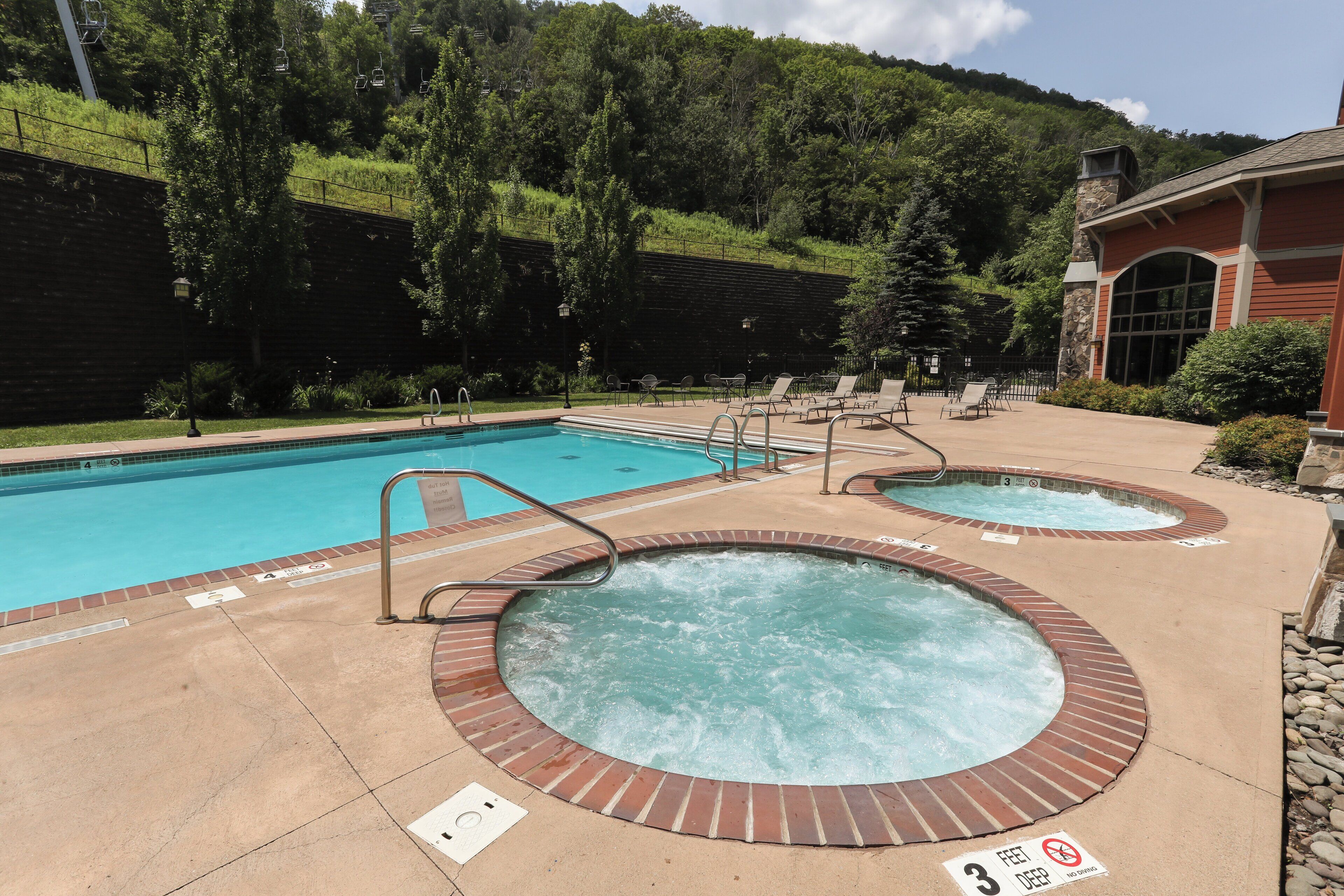 A pool area with a standard pool and two hot tubs, surrounded by lounge chairs and a building with a stone facade.