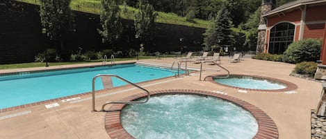 A pool area with a standard pool and two hot tubs, surrounded by lounge chairs and a building with a stone facade.