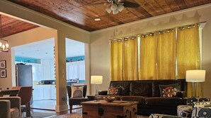 A spacious living room with a wooden ceiling, a sofa set, a patterned armchair, a wooden chest, and a dining area with a kitchen in the background. - Calming Seaside Sanctuary, pet friendly 2-blocks from beach (Galveston)