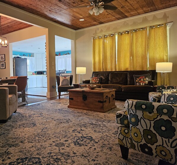A spacious living room with a wooden ceiling, a sofa set, a patterned armchair, a wooden chest, and a dining area with a kitchen in the background.