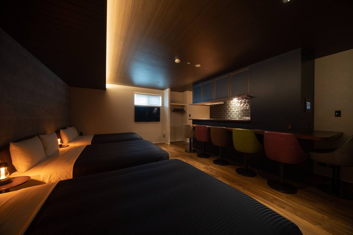 Una habitación de hotel moderna con una cama grande, una kitchenette pequeña y una zona de bar con sillas coloridas.