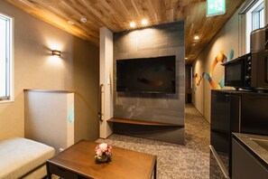 A modern hotel room with a wooden ceiling, a flat-screen TV mounted on a stone wall, a kitchenette with black appliances, and a small wooden table with a vase of flowers. - Grand Base Shianbashi (Nagasaki)