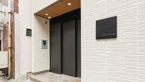 A building entrance with a set of steps leading to a door, a sign on the wall, and a security camera. - Grand Base Shianbashi (Nagasaki)