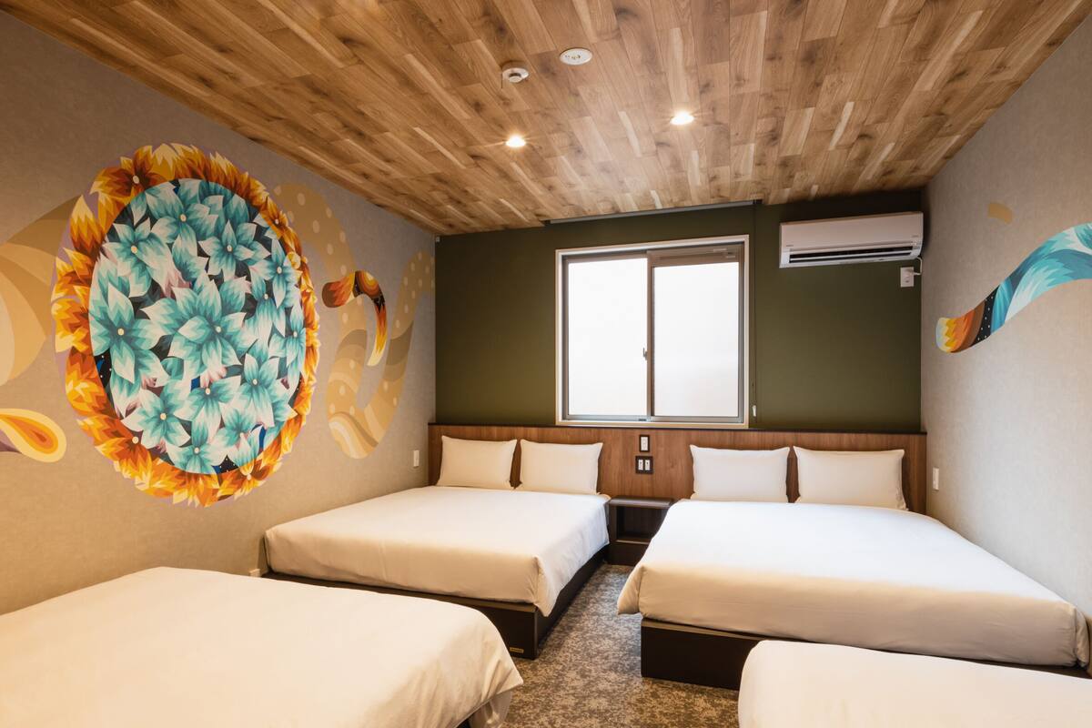 Une chambre d’hôtel avec deux lits, un plafond en bois, une grande fresque murale à motifs floraux et une fenêtre équipée d’un appareil de climatisation.