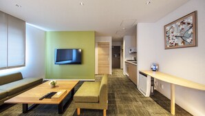 A modern hotel room with a sofa, a wooden coffee table, a bench, a TV mounted on a green wall, a kitchen area, and a painting on the wall. - Grand Base Okayama Ekimae (Okayama)