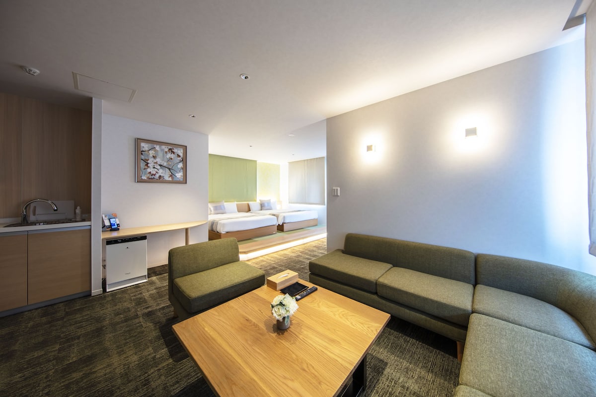 A modern hotel room with a sofa, a wooden coffee table, a kitchen area, and a bed with a green wall.