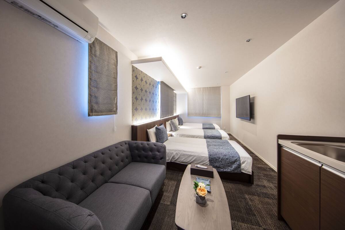 A hotel room with a grey sofa, two single beds, a TV, and a small kitchenette.