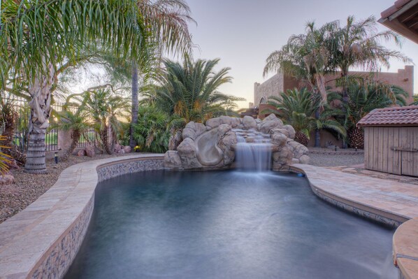 A swimming pool with a waterfall feature, surrounded by palm trees and a stone wall.