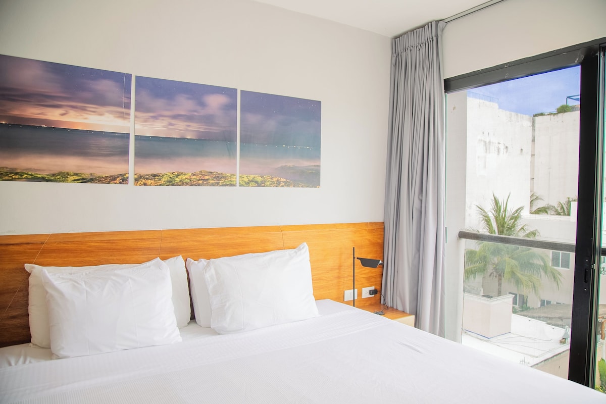 A hotel room with a wooden headboard, a large bed with white bedding, a nightstand, a lamp, and a view of a cityscape through a window.