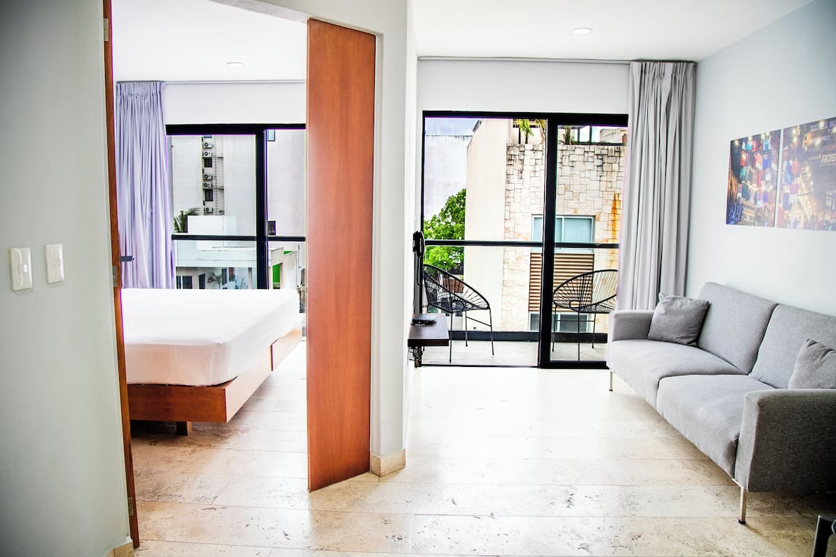 A modern hotel room with a bed, a sofa, a dining area with a table and chairs, and a balcony with a view of the outside.