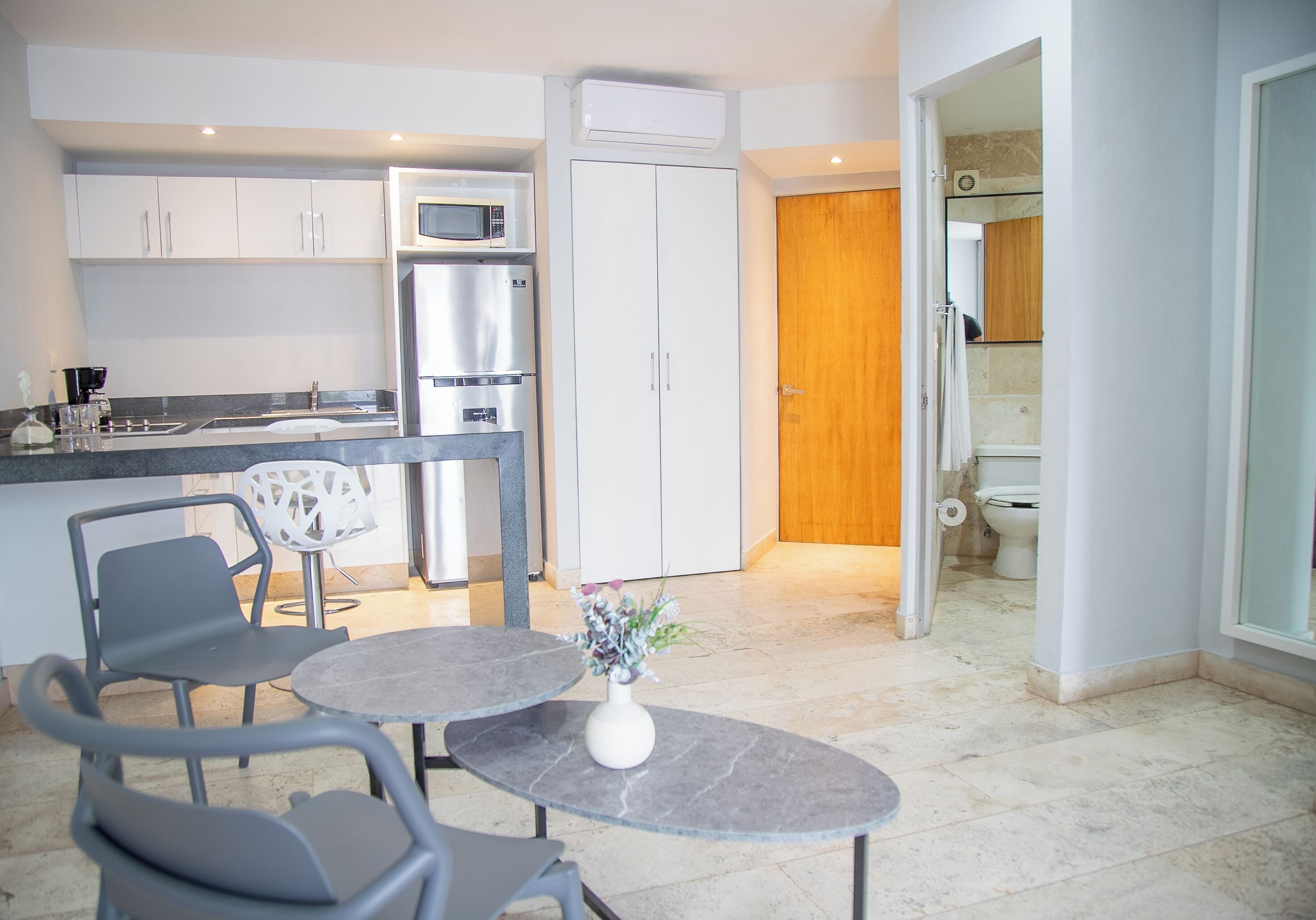 Une cuisine moderne avec un coin repas, un réfrigérateur, un four à micro-ondes et une porte en bois donnant accès à la salle de bain.