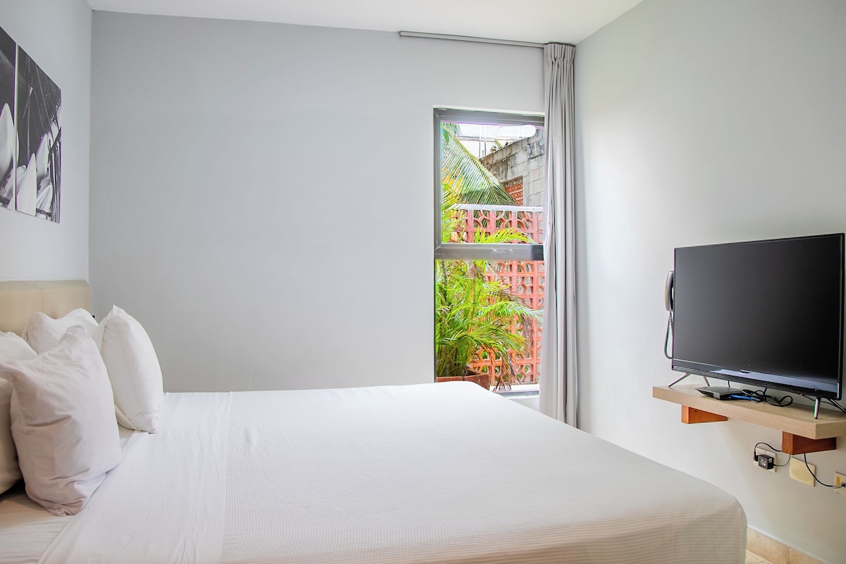A hotel room with a bed, a flat-screen TV, a window with a view of a red fence and greenery, and a wall-mounted shelf.