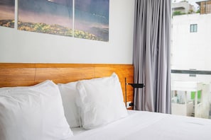 A hotel room with a wooden headboard, white bedding, a nightstand with a lamp, and a view of buildings through a window.