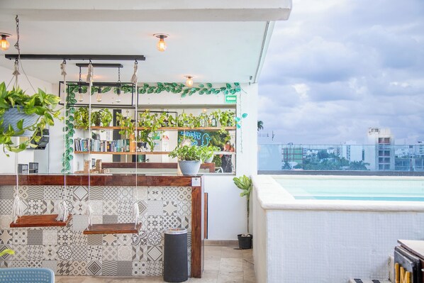 Un bar sur le toit offrant une vue sur la piscine, agrémenté de plantes suspendues et d’un mur recouvert de carreaux aux motifs originaux.