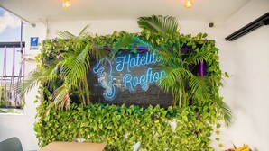 Ein Hotel-Empfangsbereich mit einer Neonreklame, Zimmerpflanzen, einem Empfangstresen und einer Obstschale.