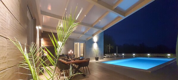 An outdoor pool area with a dining table and chairs, surrounded by plants and evening lighting. - Brand-new, luxury ***** villa with a heated pool, just 2 km from the ocean (Rakalj)