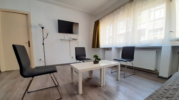 A modern room with two black chairs, a white table, a small potted plant, a flat-screen TV mounted on the wall, and a window with sheer curtains. - Central Apartment Bucharest (București)